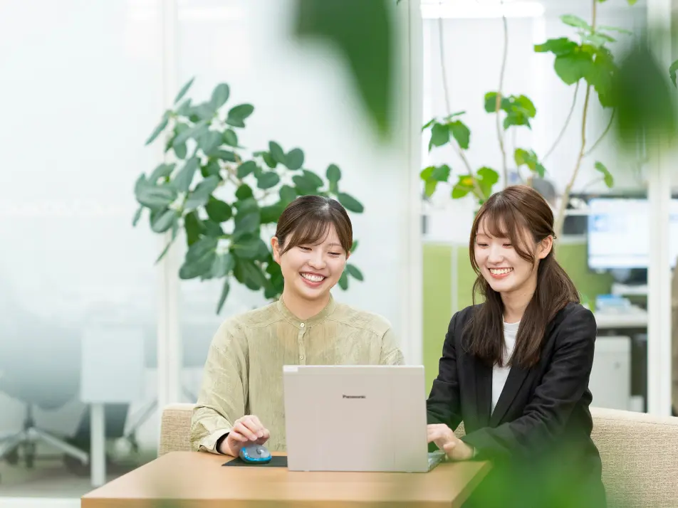 植物が並ぶ明るいオフィスでパソコンを見ながら笑顔で話す女性社員2名